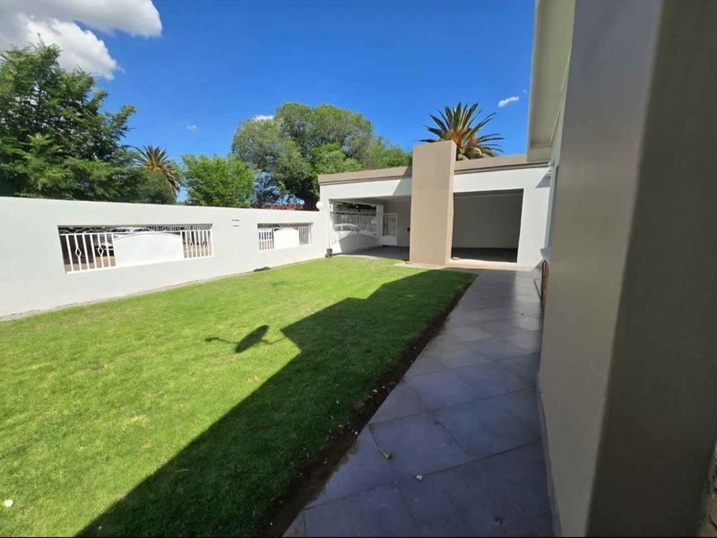 A well-maintained rear garden featuring a tiled patio, manicured lawn, and a secure boundary wall with integrated gate access to a covered parking area.