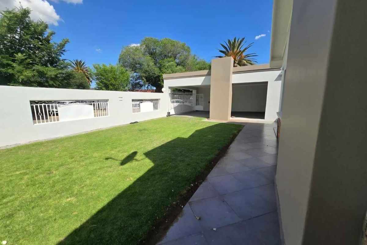 A well-maintained rear garden featuring a tiled patio, manicured lawn, and a secure boundary wall with integrated gate access to a covered parking area.