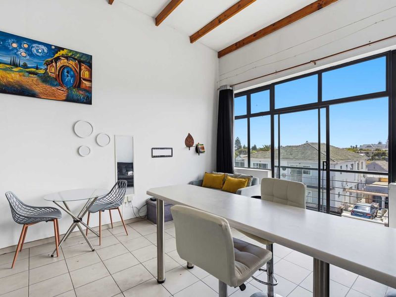 Bright open-plan living area featuring exposed timber ceiling beams, large windows providing natural light, and direct access to a balcony.