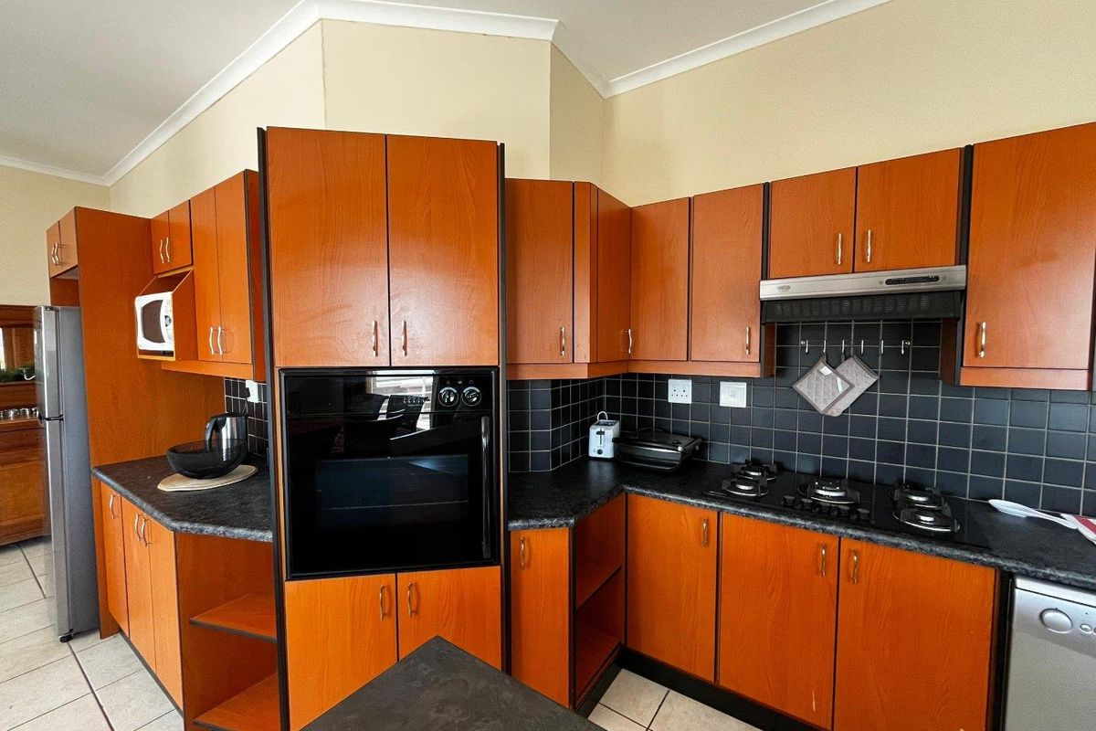A functional kitchen featuring wood-finish cabinetry, black tiled backsplash, built-in oven, gas hob, and extractor fan with tiled flooring.