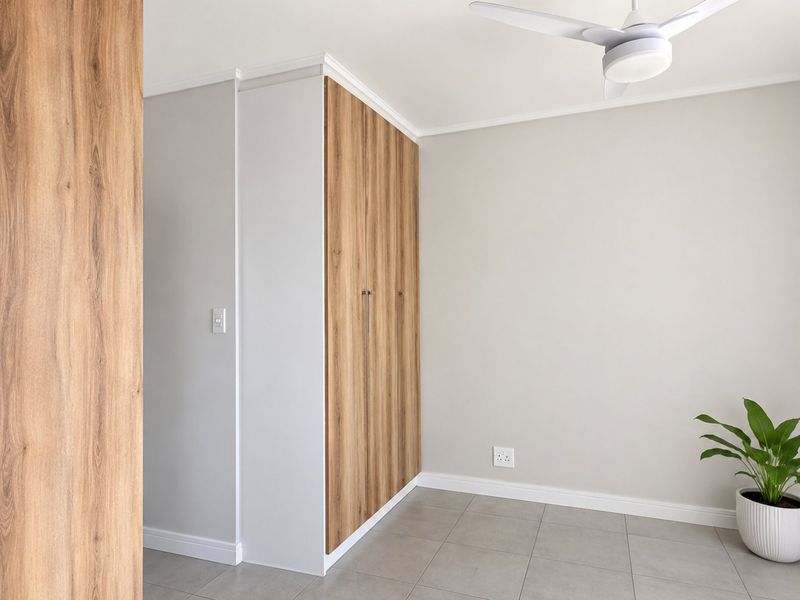 A clean, modern bedroom featuring neutral tones, tiled flooring, a ceiling fan, and a sleek built-in wardrobe with wood-grain finish.