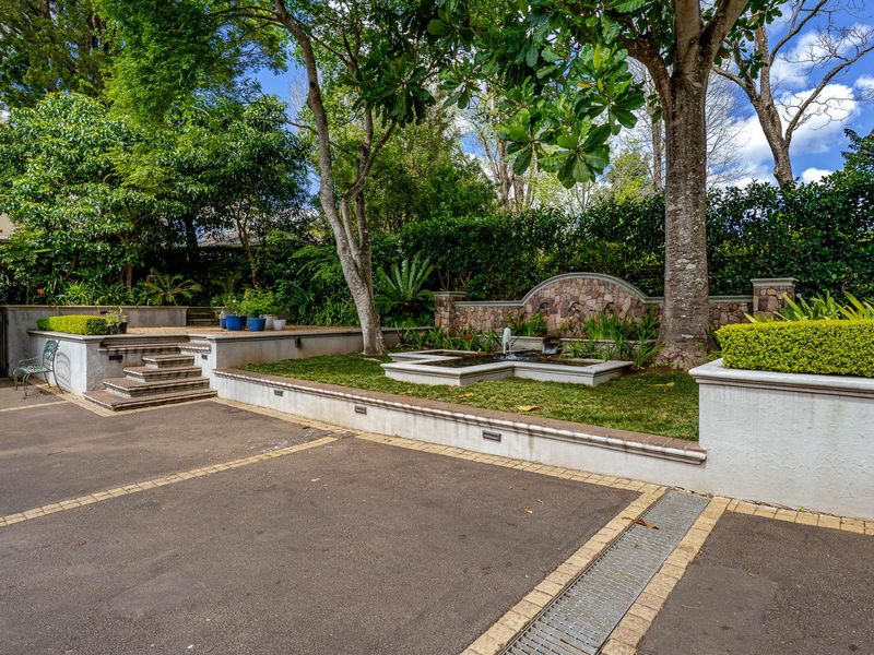 A well-maintained garden area featuring a stone retaining wall, a central water feature, paved driveway, and mature trees.