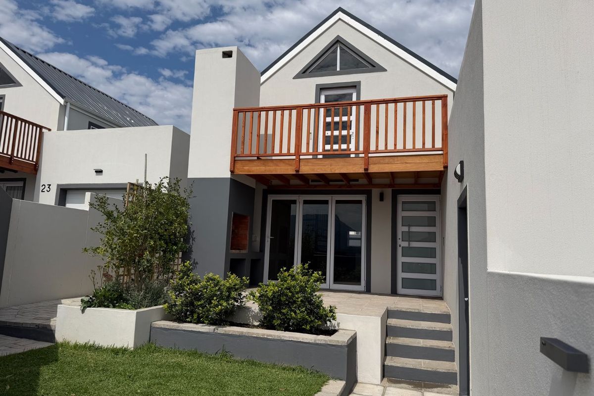 Rear exterior view featuring a paved patio, built-in braai, raised garden beds, and an upper-level wooden balcony with glass sliding door access.
