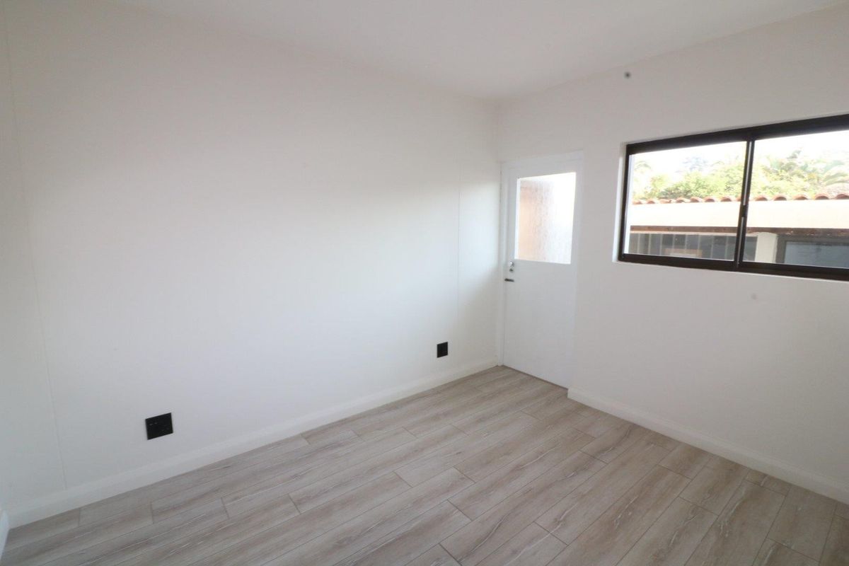 A bright, empty bedroom featuring modern light-toned laminate flooring, a sliding window, and a private external access door.