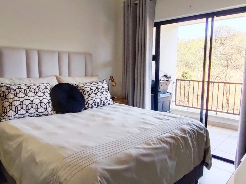 A well-lit bedroom featuring neutral tones, tiled flooring, and a sliding glass door providing access to a private balcony with outdoor views.