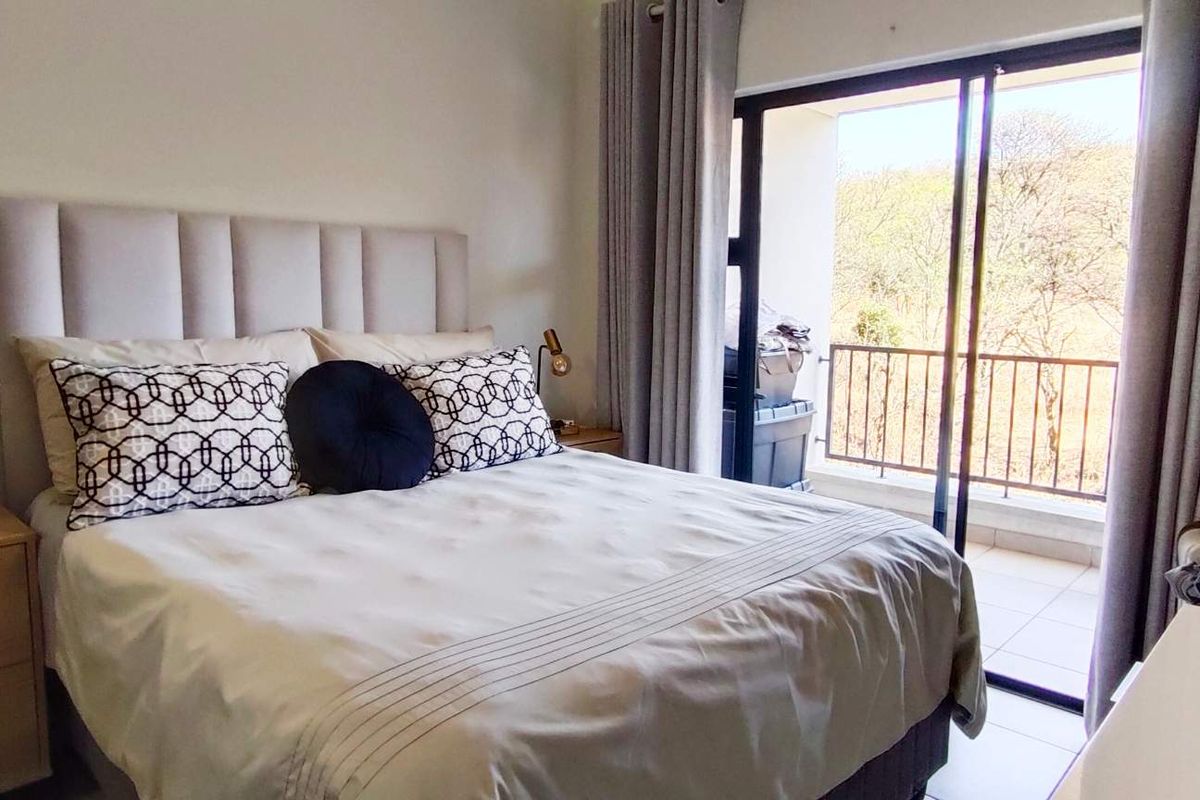 A well-lit bedroom featuring neutral tones, tiled flooring, and a sliding glass door providing access to a private balcony with outdoor views.