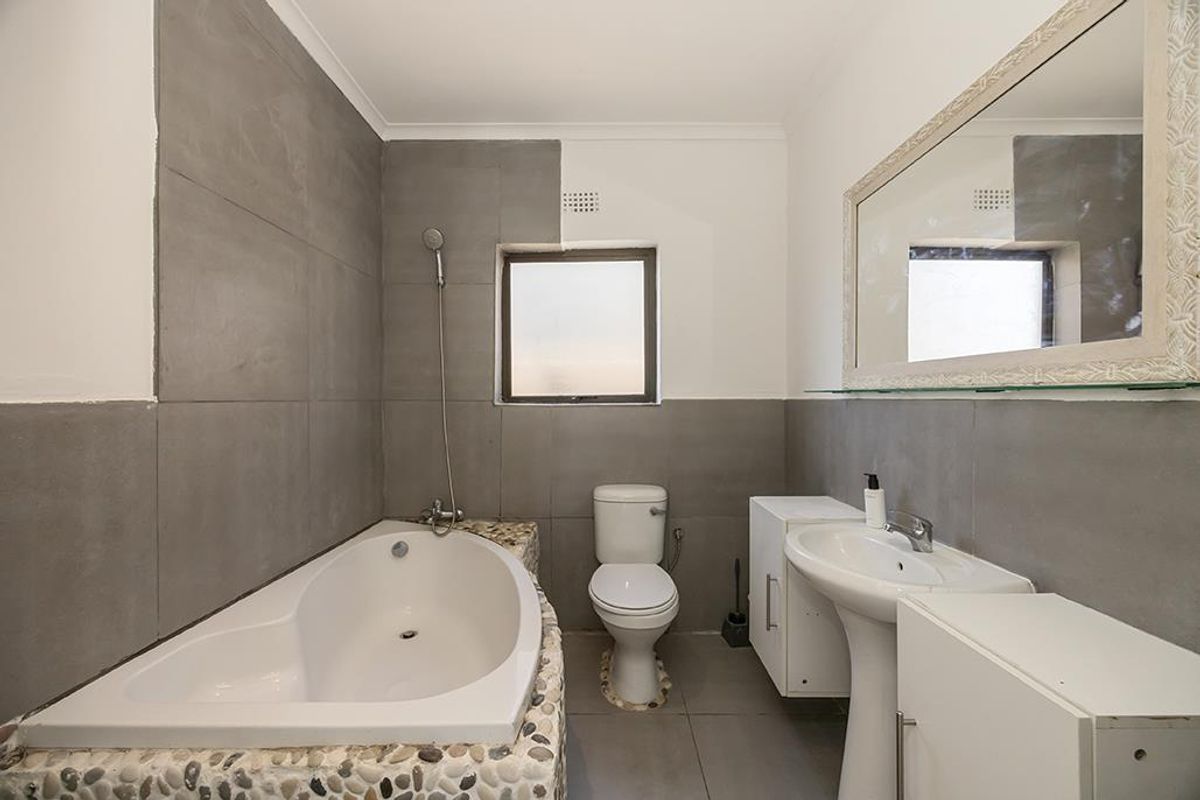 A functional bathroom featuring a built-in bathtub with pebble-tiled surround, a pedestal basin, and a standard toilet with grey wall tiling.