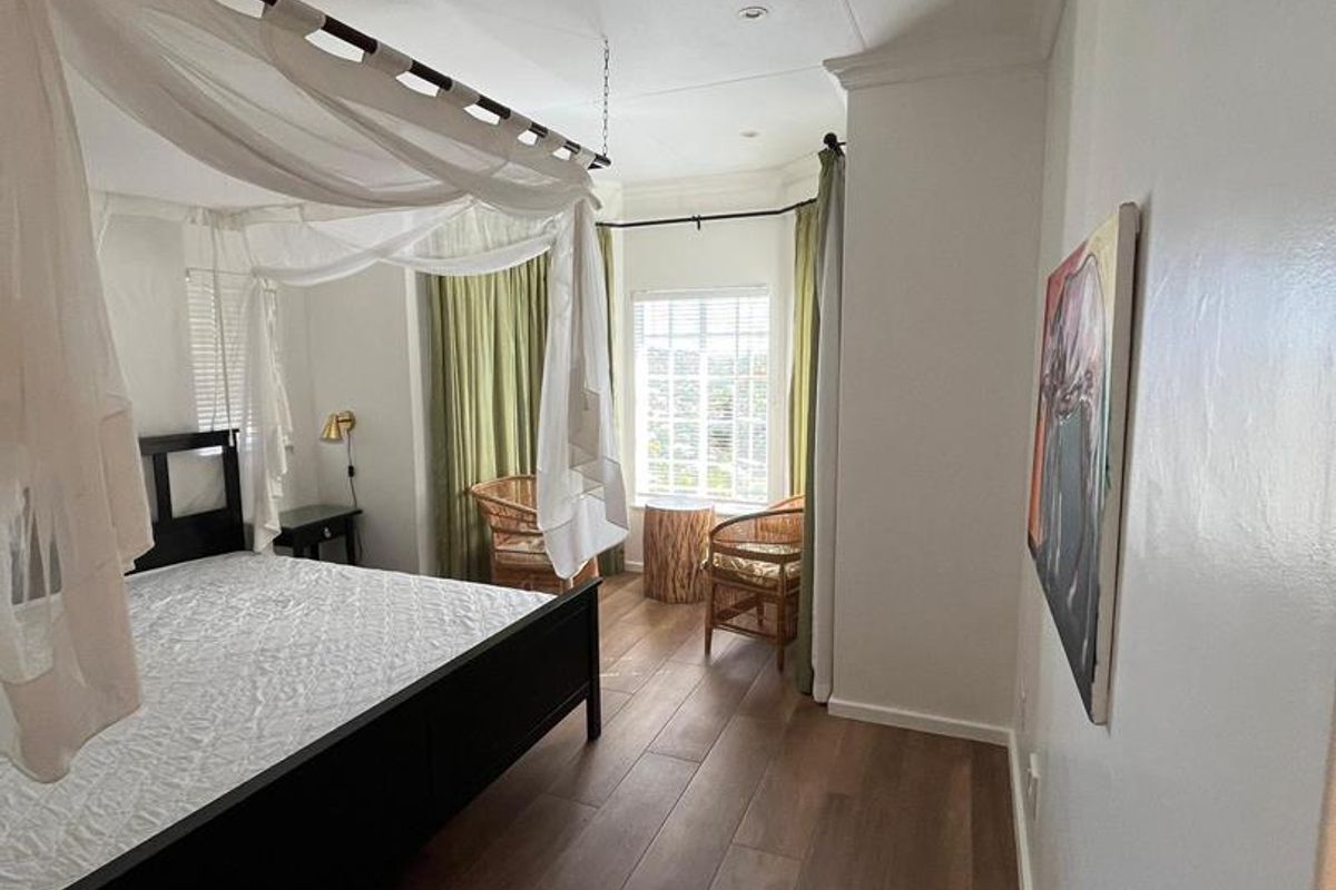 A bright bedroom featuring wood-look flooring, recessed ceiling lighting, and a large window with curtains, creating a comfortable and inviting sleeping space.