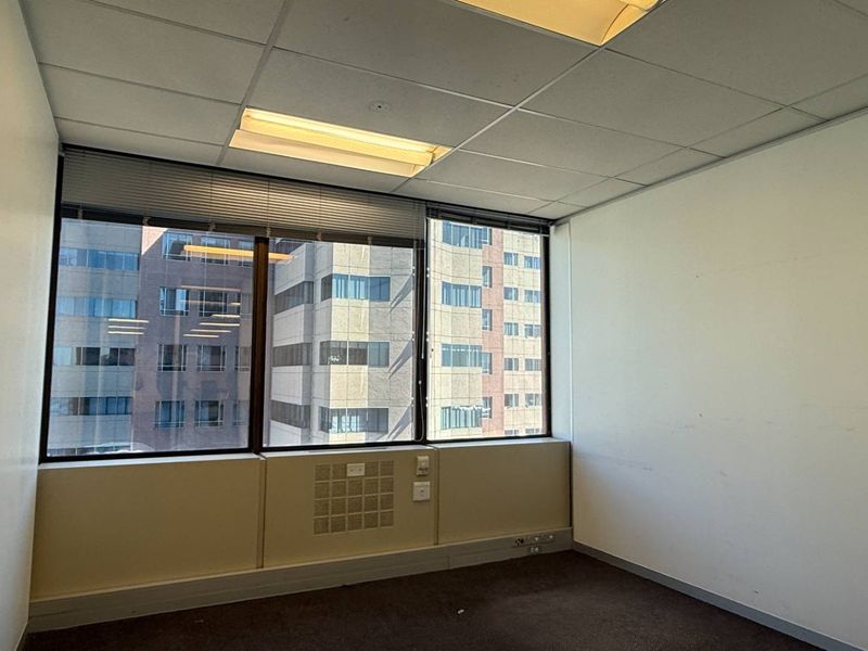 A vacant office space featuring a suspended ceiling, recessed lighting, large windows with blinds, and carpet flooring, offering a functional workspace with city views.