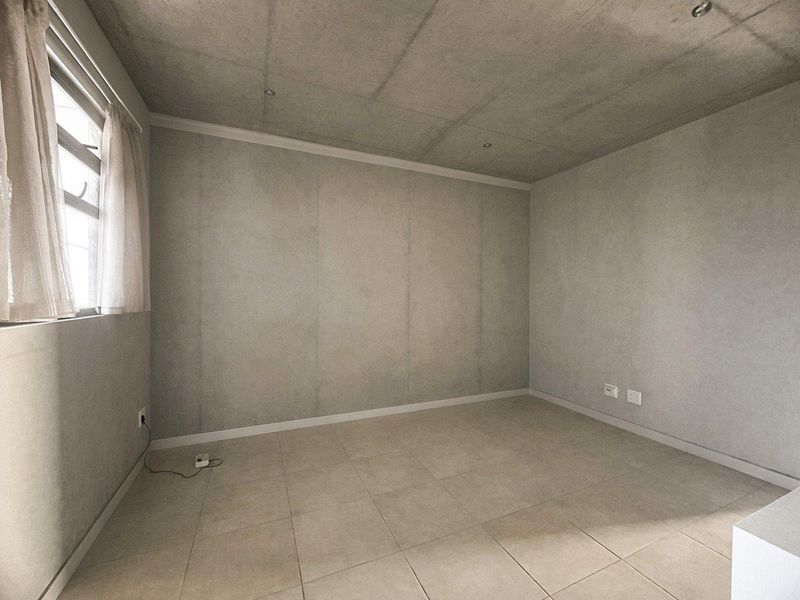 An empty bedroom featuring an exposed concrete ceiling, neutral tiled flooring, and a window, offering a clean, modern blank canvas.