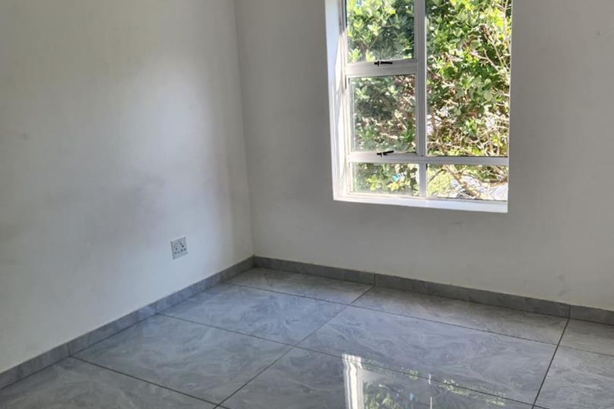 A bright, empty bedroom featuring modern grey tiled flooring, a single pendant light, and a window with a view of greenery.