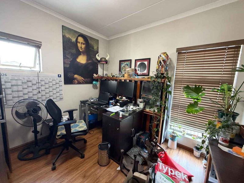 A functional home office space featuring laminate flooring, natural light through venetian blinds, and a top-hung window.