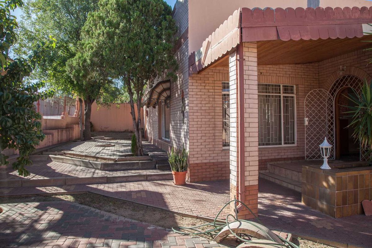 A view of the rear exterior featuring a brick facade, covered patio area, and paved garden space with mature trees.