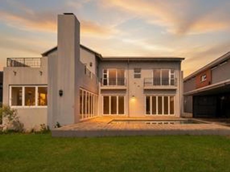 Rear view of a modern two-story house featuring a swimming pool, wooden decking, balconies, and large sliding glass doors at sunset.