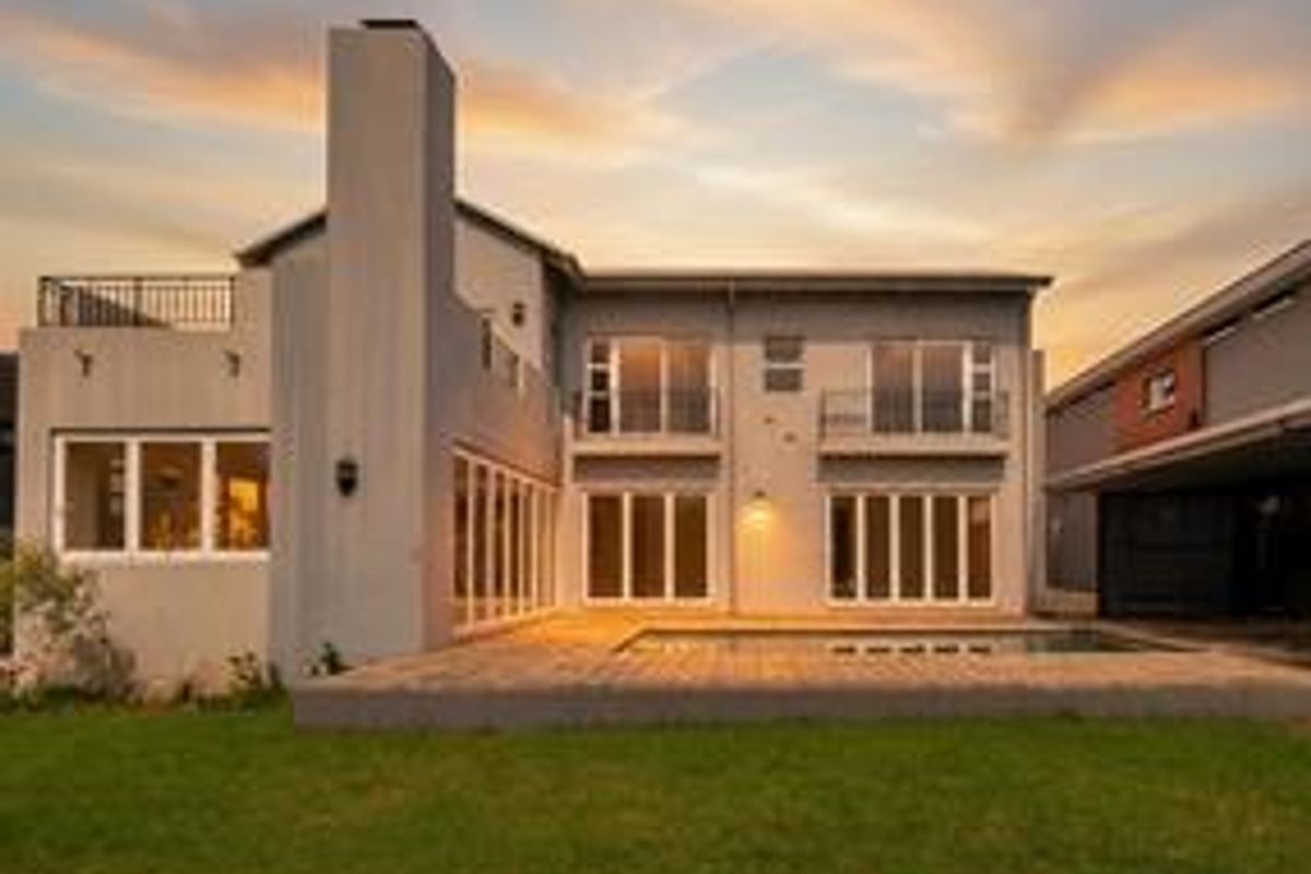 Rear view of a modern two-story house featuring a swimming pool, wooden decking, balconies, and large sliding glass doors at sunset.