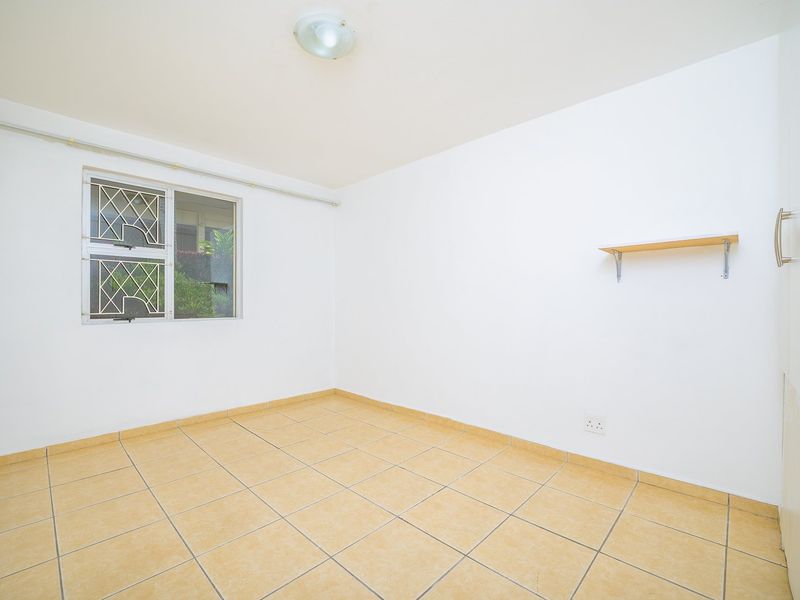 A bright, empty bedroom featuring tiled flooring, a built-in cupboard, and a window fitted with security bars.