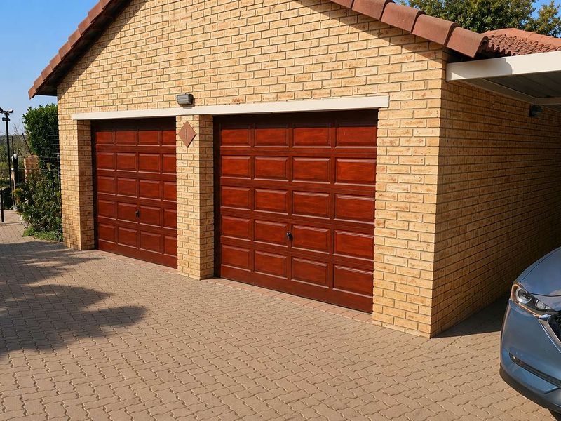 A well-maintained double garage featuring classic face brick construction, timber-look sectional doors, and an adjacent paved carport area.