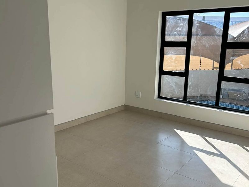 A bright, empty bedroom featuring neutral tiled flooring and a modern black aluminium window frame, offering a clean and versatile space.