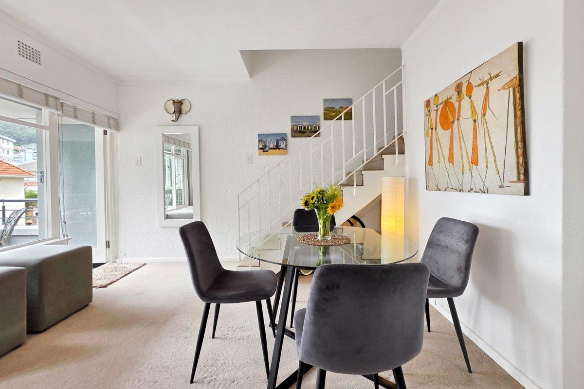 A bright living and dining area featuring carpeted flooring, a staircase with a white metal railing, and access to an outdoor balcony.