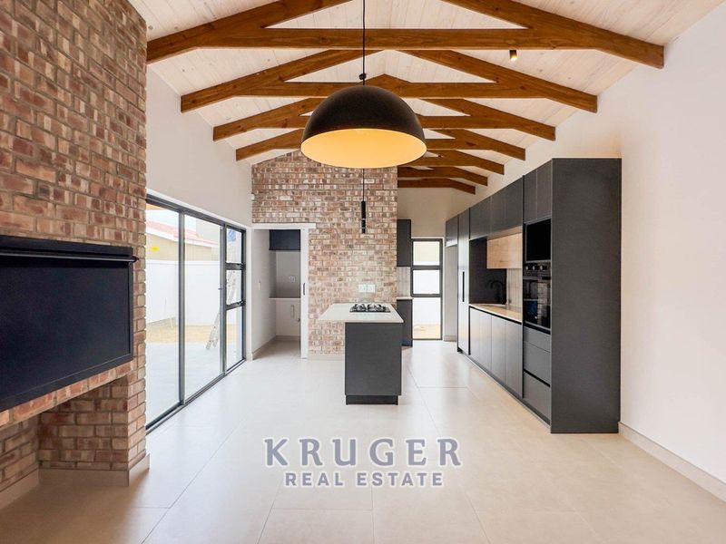 Modern open-plan living and kitchen area featuring exposed timber trusses, a brick feature wall, built-in fireplace, and sleek contemporary cabinetry.