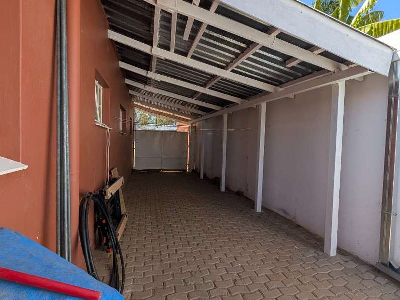 A covered outdoor walkway or side patio featuring paved flooring and a corrugated roof structure, providing a functional transition space along the side of the house.