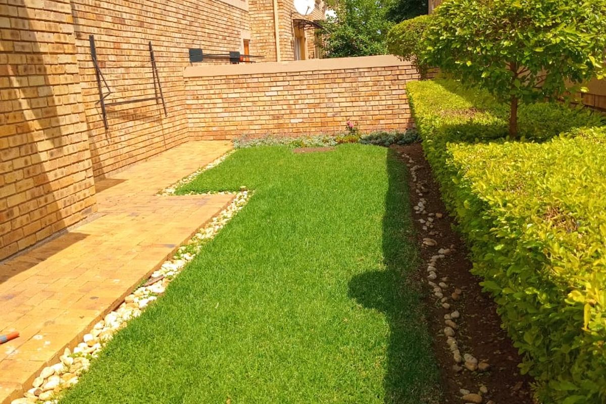 A private rear garden area featuring a well-maintained lawn, paved walkway, and established hedging, set against a brick apartment building facade.