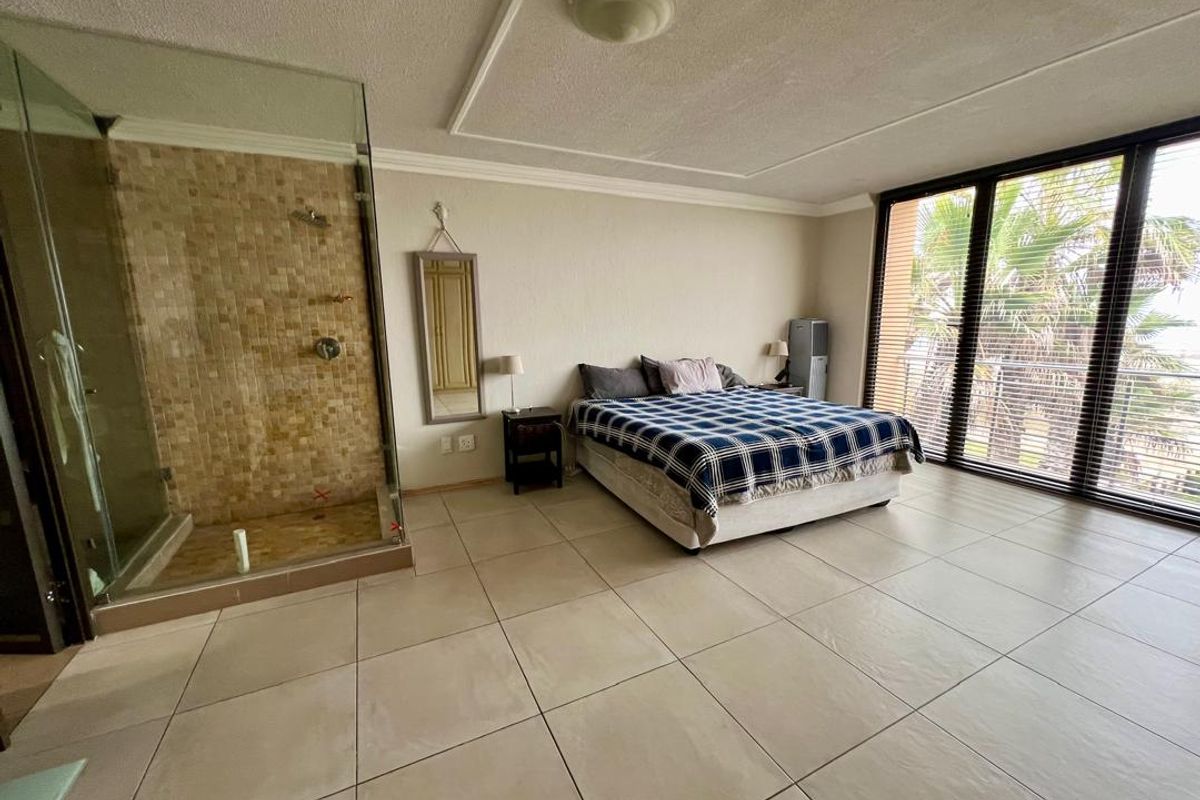 Spacious bedroom featuring an open-plan en-suite shower with mosaic tiling, large windows, and neutral floor tiles.