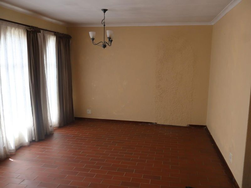 An empty living room featuring terracotta tile flooring, a central chandelier, and large windows with curtain rails, requiring cosmetic wall repairs.