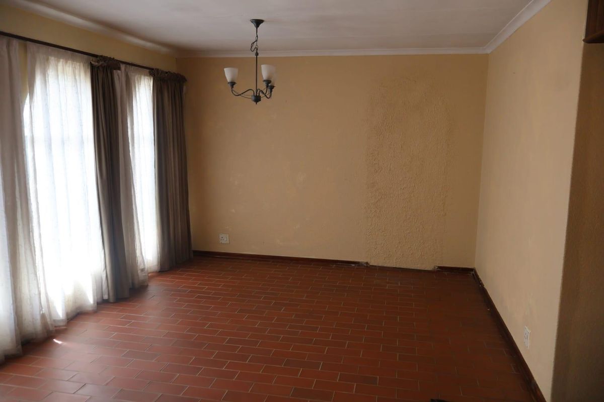 An empty living room featuring terracotta tile flooring, a central chandelier, and large windows with curtain rails, requiring cosmetic wall repairs.