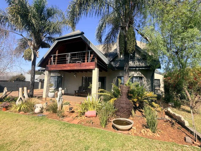Rear view of a house featuring a covered patio, an upper balcony with timber decking, stone pillars, and a well-maintained landscaped garden.
