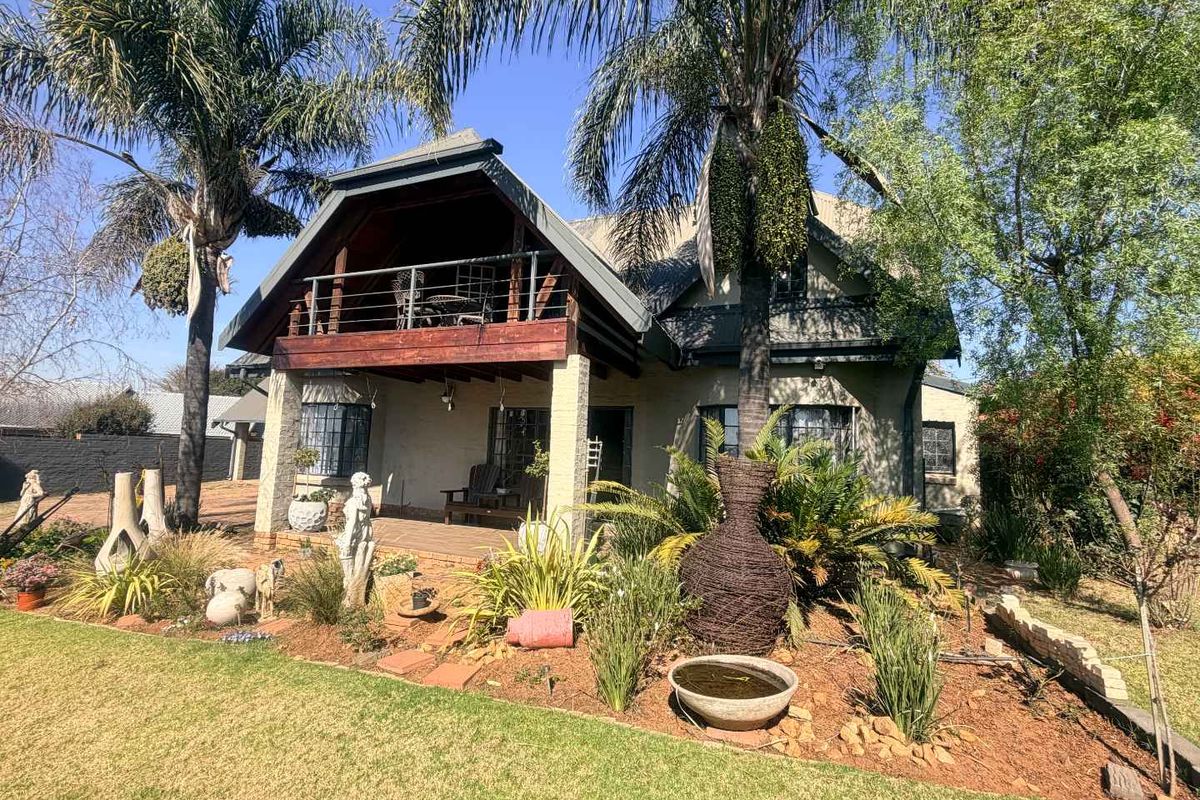 Rear view of a house featuring a covered patio, an upper balcony with timber decking, stone pillars, and a well-maintained landscaped garden.