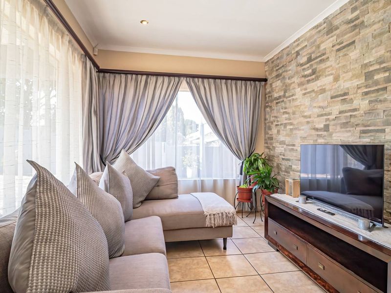 A well-maintained living room featuring a textured stone cladding feature wall, neutral tiled flooring, and large windows with curtain tracks.