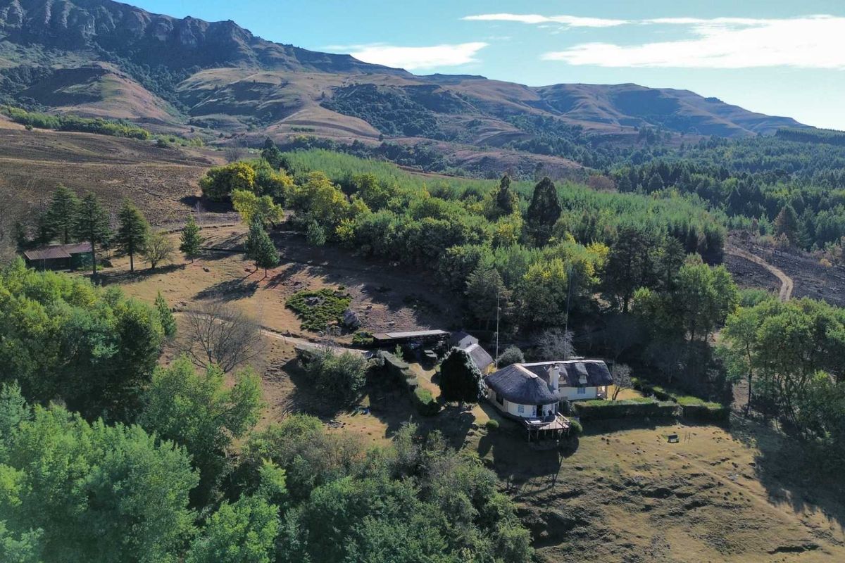 Aerial view of a rural farm property featuring a thatched-roof dwelling with an elevated deck, set against a scenic mountainous landscape.