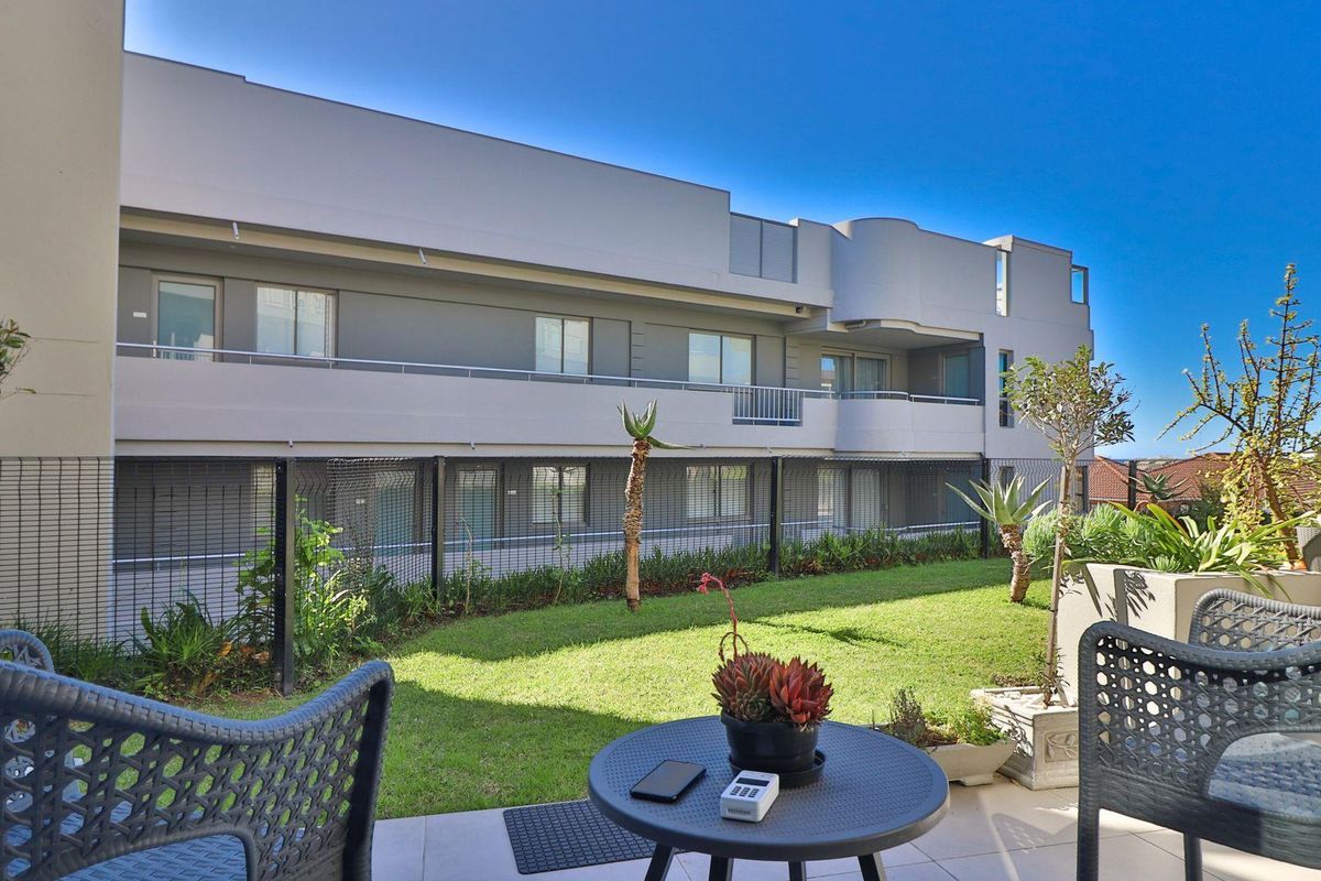 A view of the apartment building exterior featuring a private garden area and paved patio space.