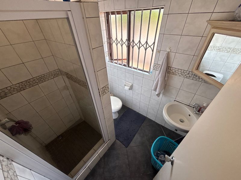 A compact bathroom featuring a shower enclosure, wall-mounted basin, and toilet, finished with tiled walls and flooring and a security-fitted window.