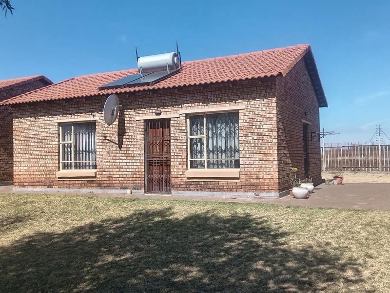 A well-maintained face brick townhouse featuring a tiled roof, solar water heating system, and secure entrance with a paved patio area.