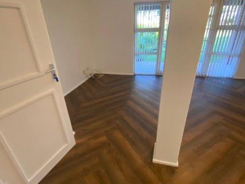 An empty living room featuring modern herringbone flooring, a structural column, and large windows with vertical blinds providing natural light.