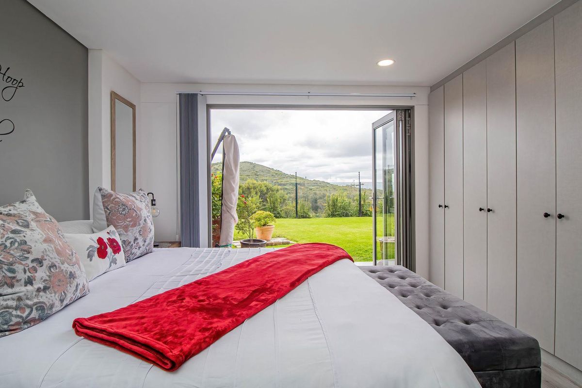 Modern bedroom featuring extensive built-in cupboards, recessed lighting, and bi-fold doors opening onto a landscaped garden with scenic hill views.