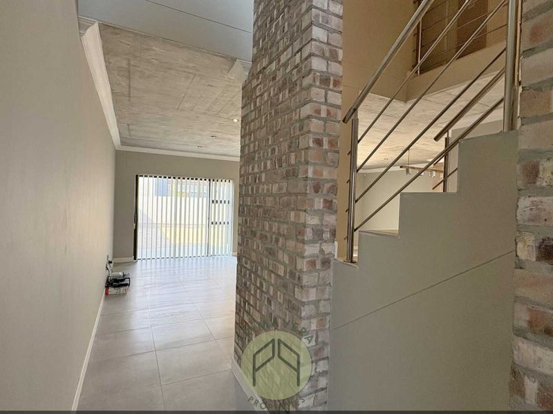 Spacious open-plan living area featuring a striking exposed brick wall, modern staircase with stainless steel balustrade, and contemporary exposed concrete ceiling finishes.