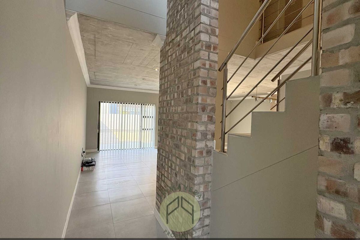 Spacious open-plan living area featuring a striking exposed brick wall, modern staircase with stainless steel balustrade, and contemporary exposed concrete ceiling finishes.