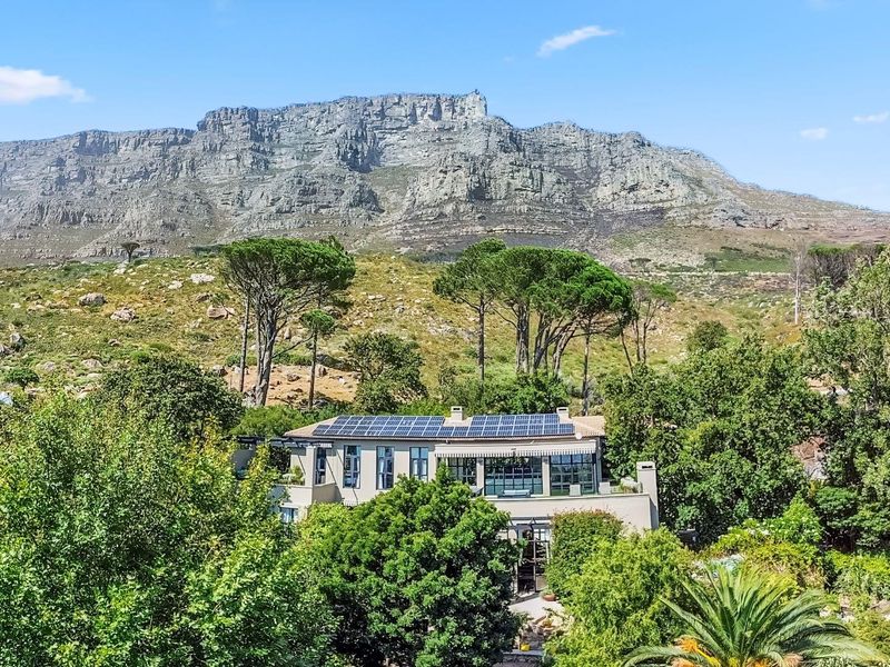 An elevated aerial view of the property featuring a large roof-mounted solar panel array, set against a dramatic mountain backdrop with lush surrounding greenery.