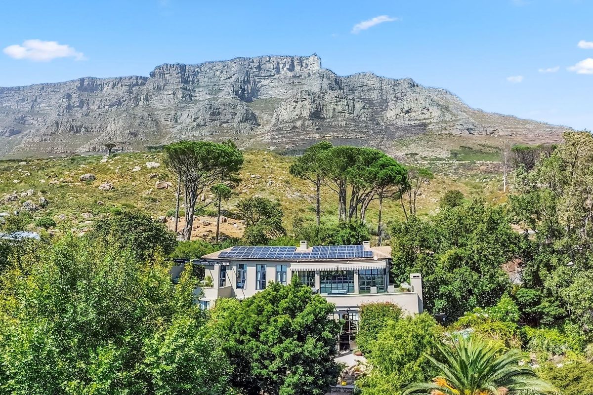 An elevated aerial view of the property featuring a large roof-mounted solar panel array, set against a dramatic mountain backdrop with lush surrounding greenery.