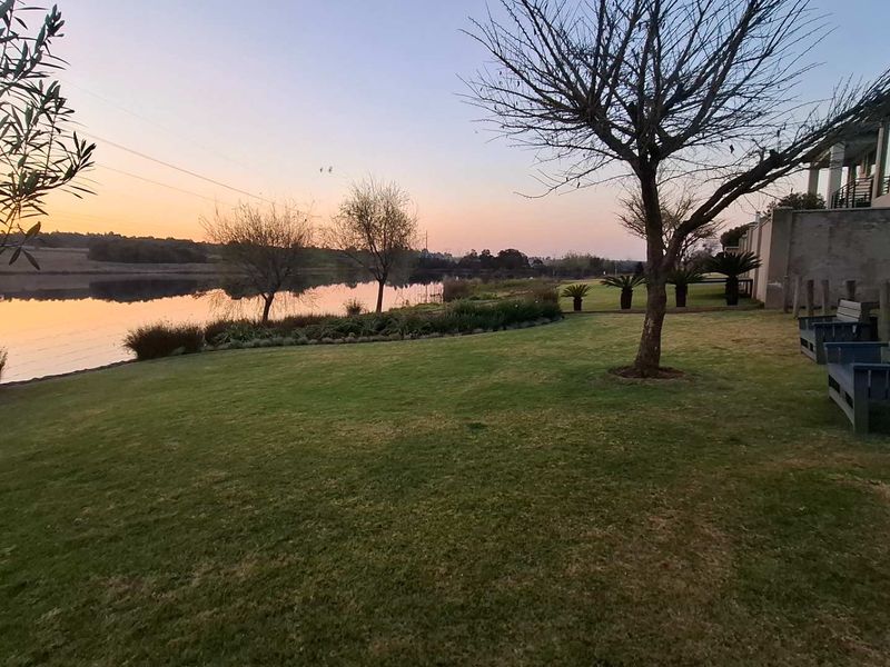 A scenic view of a tranquil waterfront area featuring a well-maintained lawn, mature trees, and a peaceful sunset backdrop.