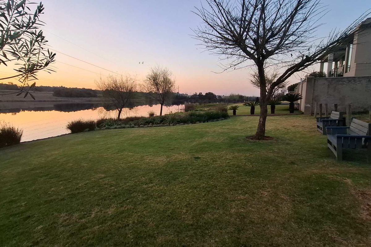 A scenic view of a tranquil waterfront area featuring a well-maintained lawn, mature trees, and a peaceful sunset backdrop.