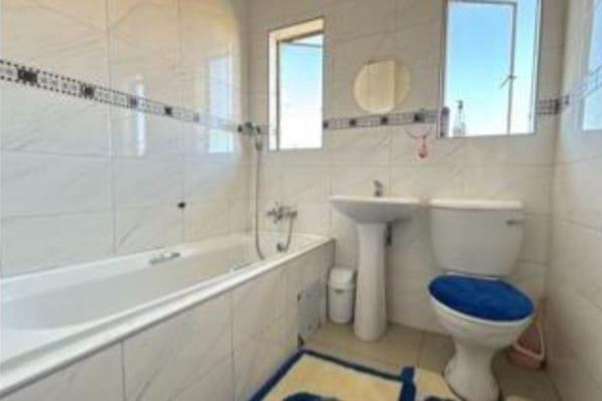 A functional bathroom featuring a white bathtub, pedestal basin, and toilet with full-height wall tiling and two windows for natural light.