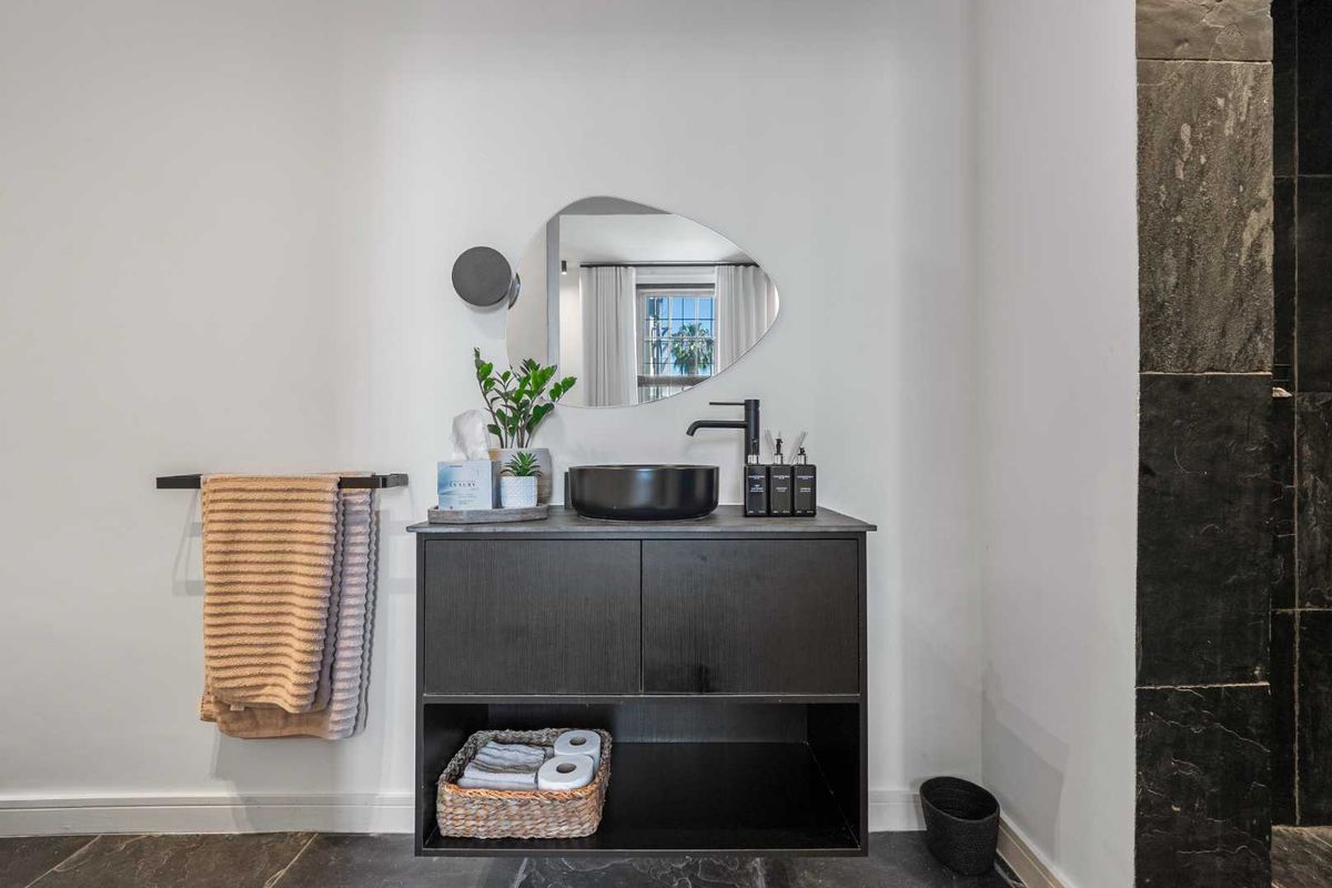 Modern bathroom vanity featuring a sleek black cabinet, vessel sink, matte black faucet, and contemporary wall mirror with stone flooring.