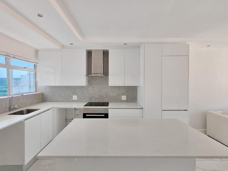 Modern kitchen featuring sleek white cabinetry, stone countertops, a central island, and integrated appliances, complemented by recessed lighting and a coastal view.