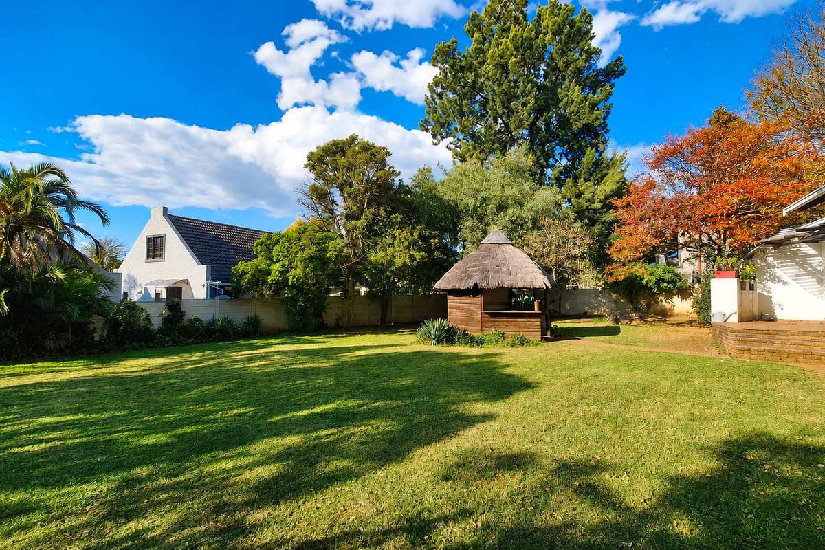 A spacious, well-maintained rear garden featuring a manicured lawn, mature trees, and a charming thatched-roof gazebo structure.