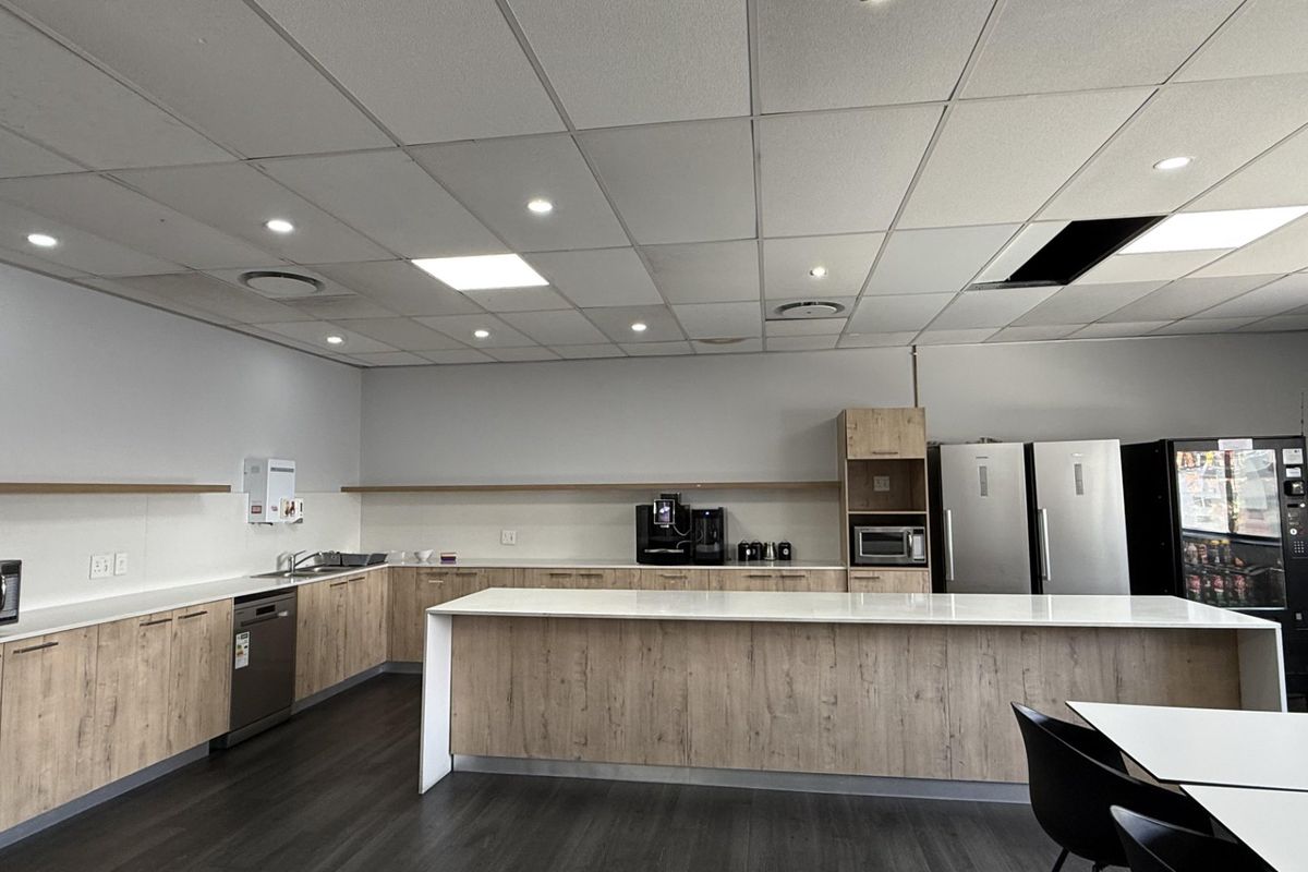 Modern office kitchen area featuring light wood cabinetry, stone countertops, integrated appliances, and a functional island workspace under a suspended ceiling.