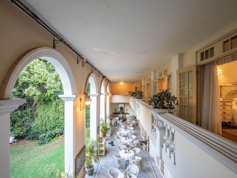 A spacious covered balcony featuring classic arched columns, a decorative balustrade, and tiled flooring, overlooking the garden area.